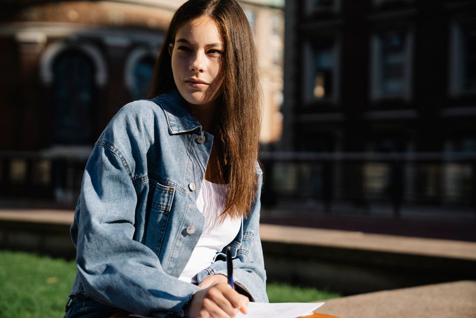 A young woman in a denim jacket studying outdoors in the sunlight, with focus and intent.