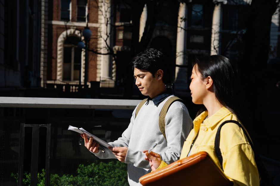 Two students walk and discuss notes outdoors on a university campus in bright daylight.