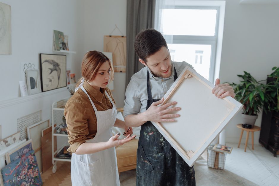 Two artists in aprons discuss art techniques with a blank canvas in a creative studio.