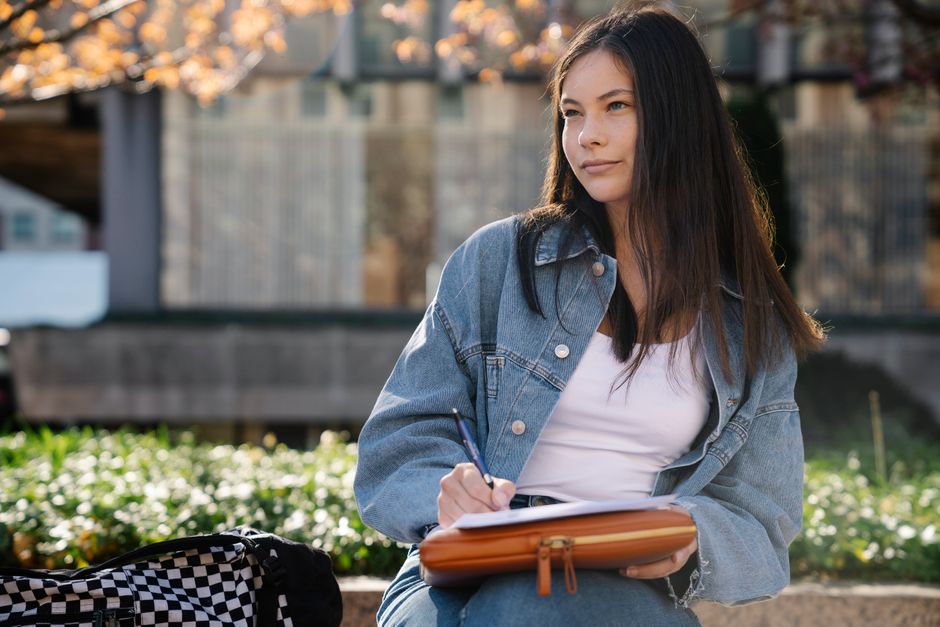 A young woman in a denim jacket writing outdoors in a university campus setting.