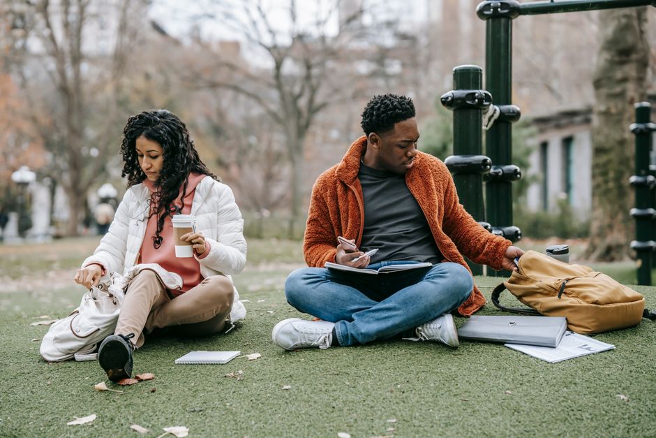 Full body of ethnic woman with takeaway coffee and black man with notebook opening rucksacks while sitting on grassy ground during studies together