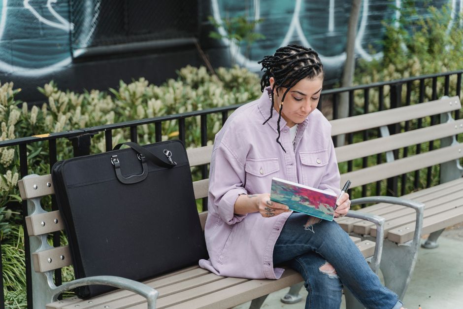 Concentrated ethnic female artist in casual clothes sitting on bench in park and drawing in sketchbook