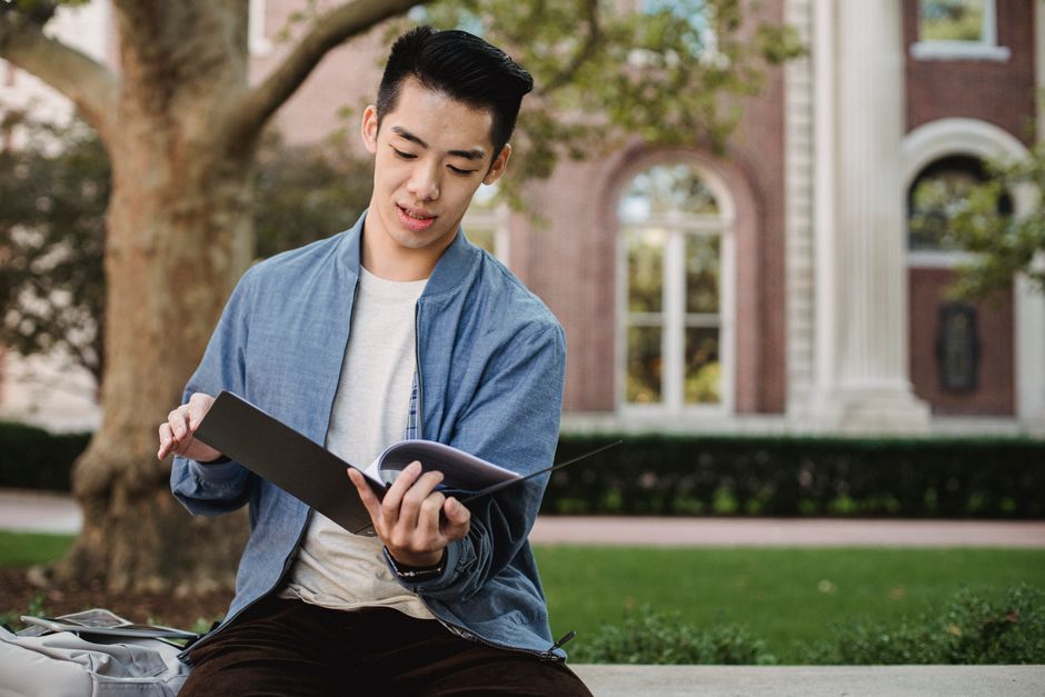 Concentrated Asian male student sitting on bench and reading information on papers in folder for homework